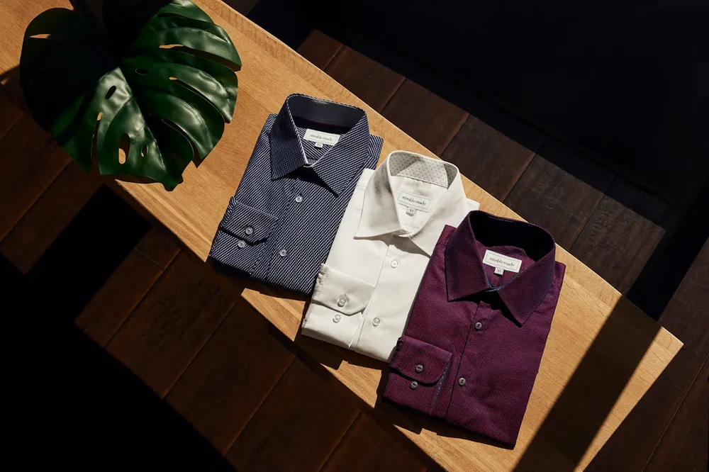 Trei cămăși împăturite — bleumarin, alb, grena — pe masă de lemn